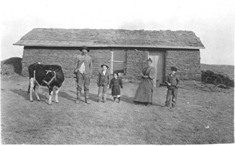 Dam och herre med tre barn och en ko framför mindre torp. Foto: Svenska Emigrantinstitutet